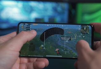 A person is holding a smartphone horizontally, playing a video game that involves a character parachuting into an open area. The screen shows various game interface elements including a compass, a mini-map, and player stats. The background has a blurred and colorful setting, suggesting a focus on the gaming experience.